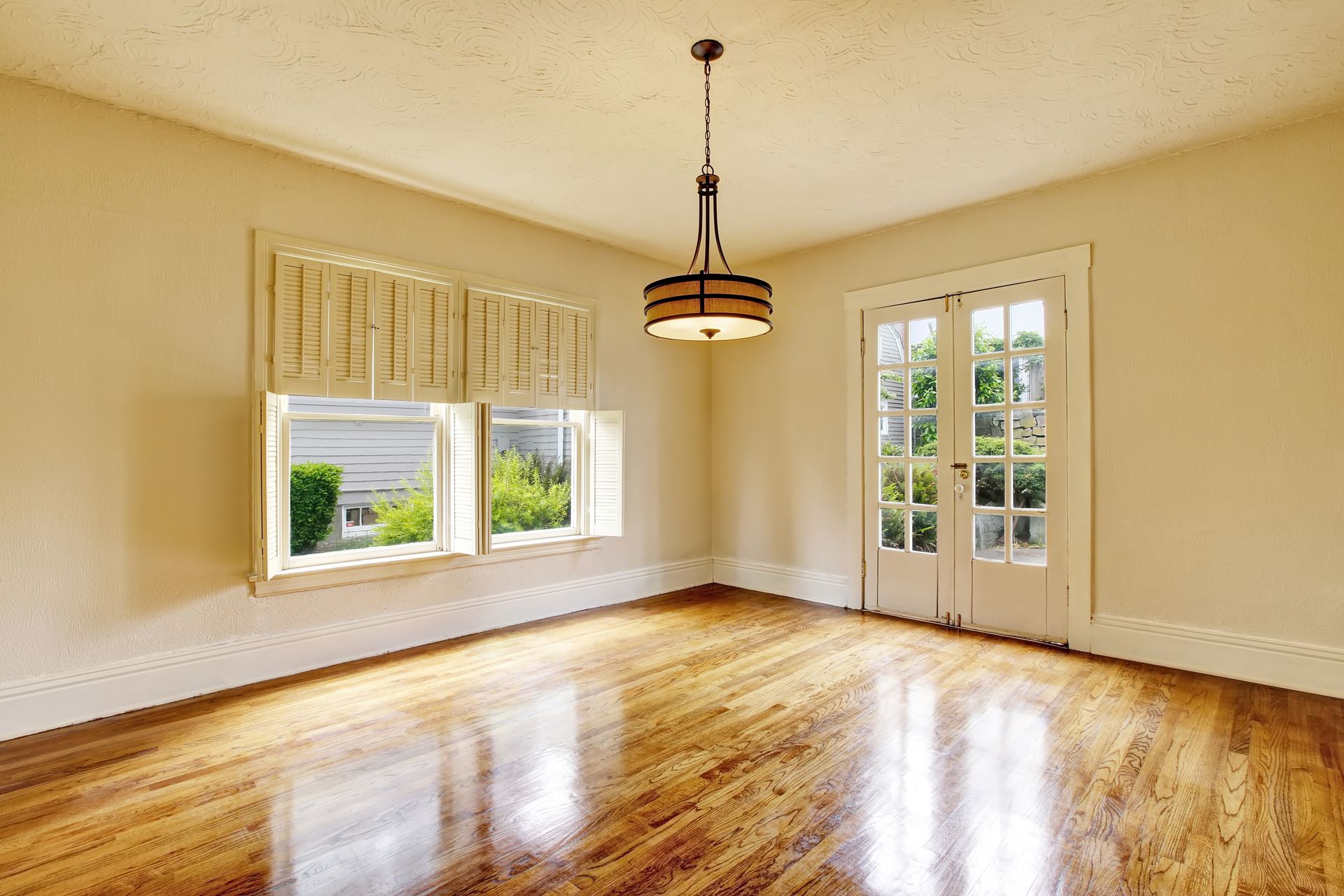An empty room with light-colored walls, polished hardwood floors, a tiered pendant light, and French doors to a garden.