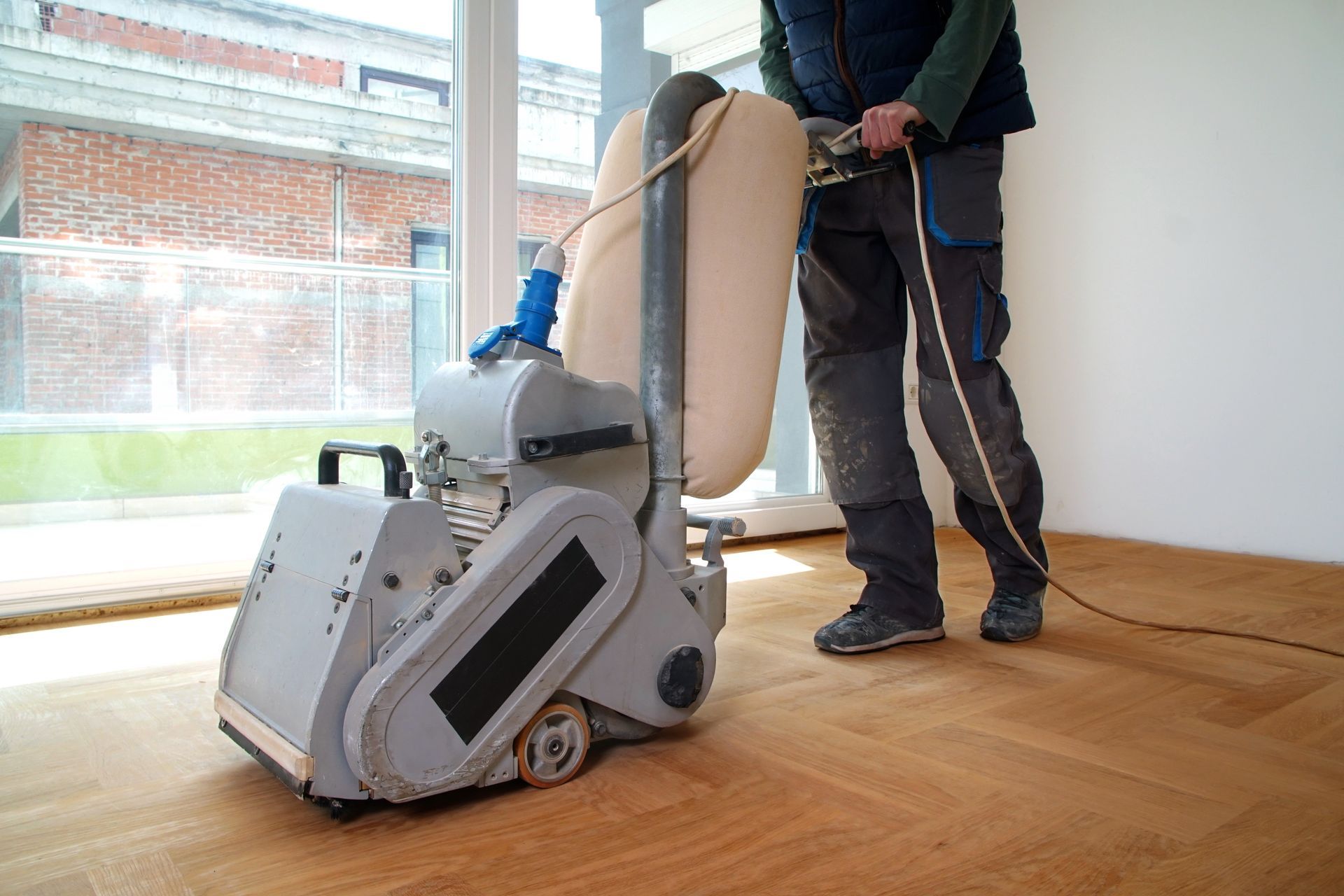A worker uses a large, grey electric floor sander to refinish wooden parquet flooring in a room with large windows.