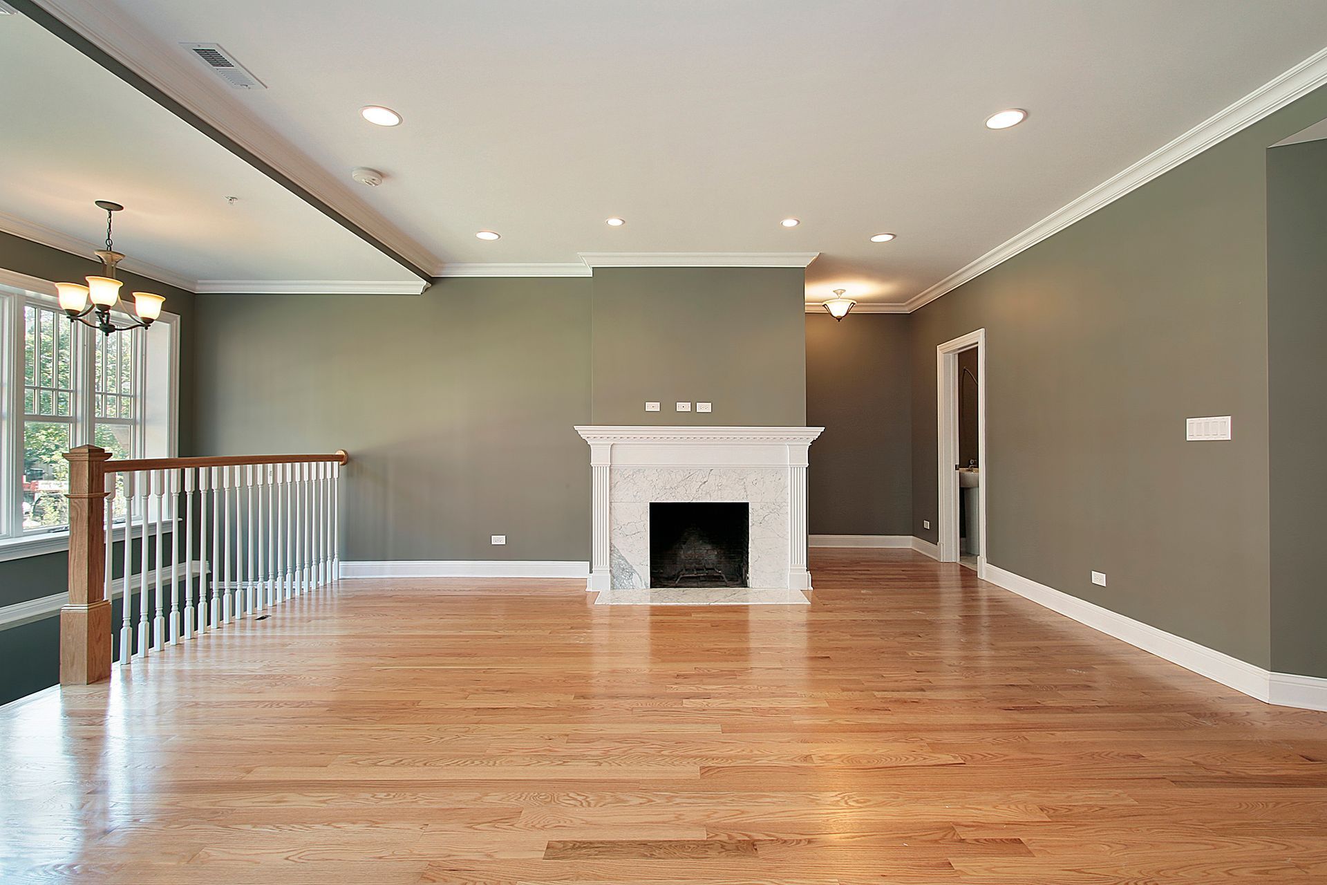 A spacious living room featuring hardwood floors, olive-gray walls, a white fireplace, and a white staircase railing.