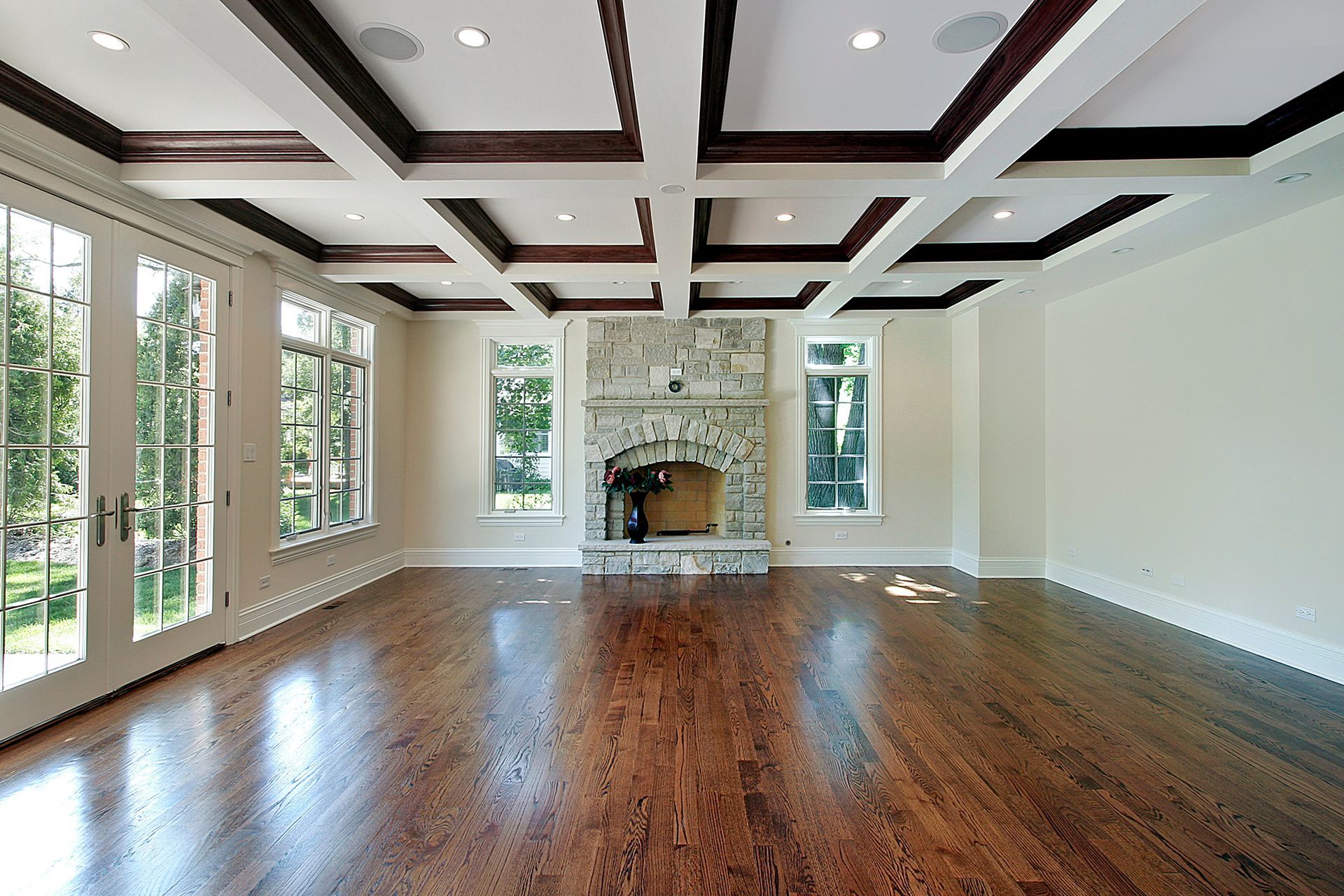 An empty living room with dark hardwood floors, a coffered ceiling, a stone fireplace, and French doors.
