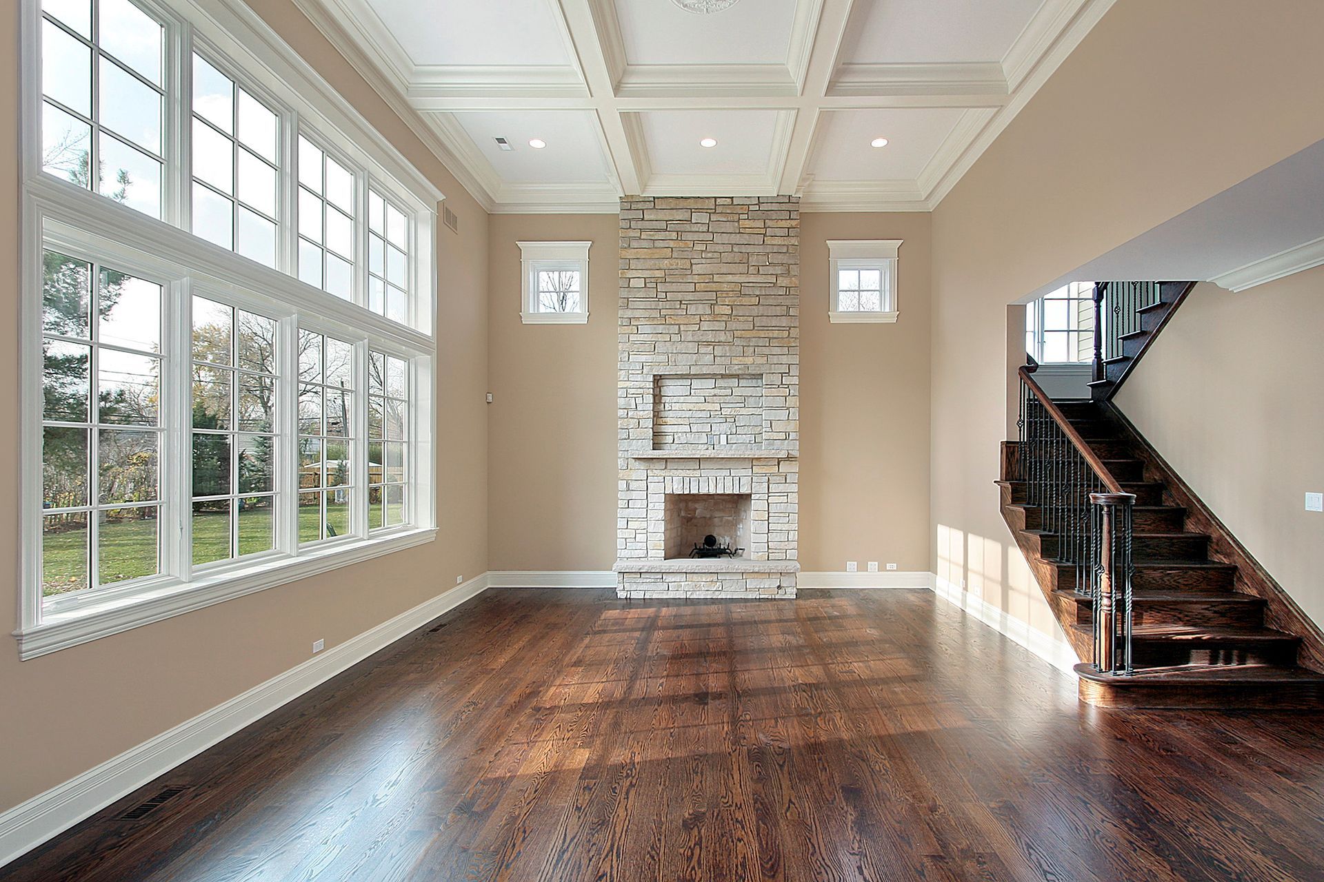 An empty room with dark hardwood floors, tan walls, a floor-to-ceiling stone fireplace, and large windows.