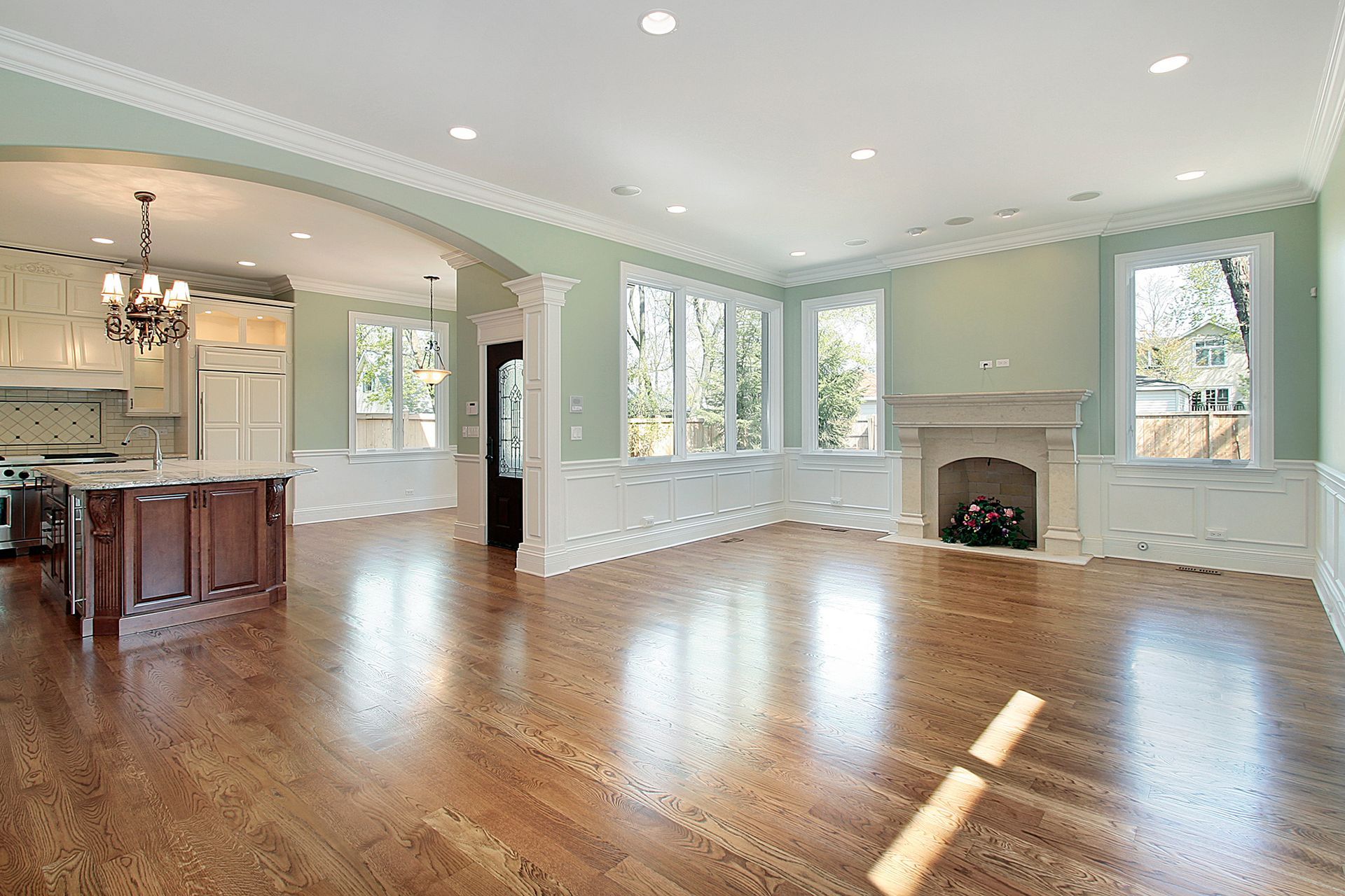 A spacious, bright open-plan room with hardwood floors, light green walls, white crown molding, and a stone fireplace.