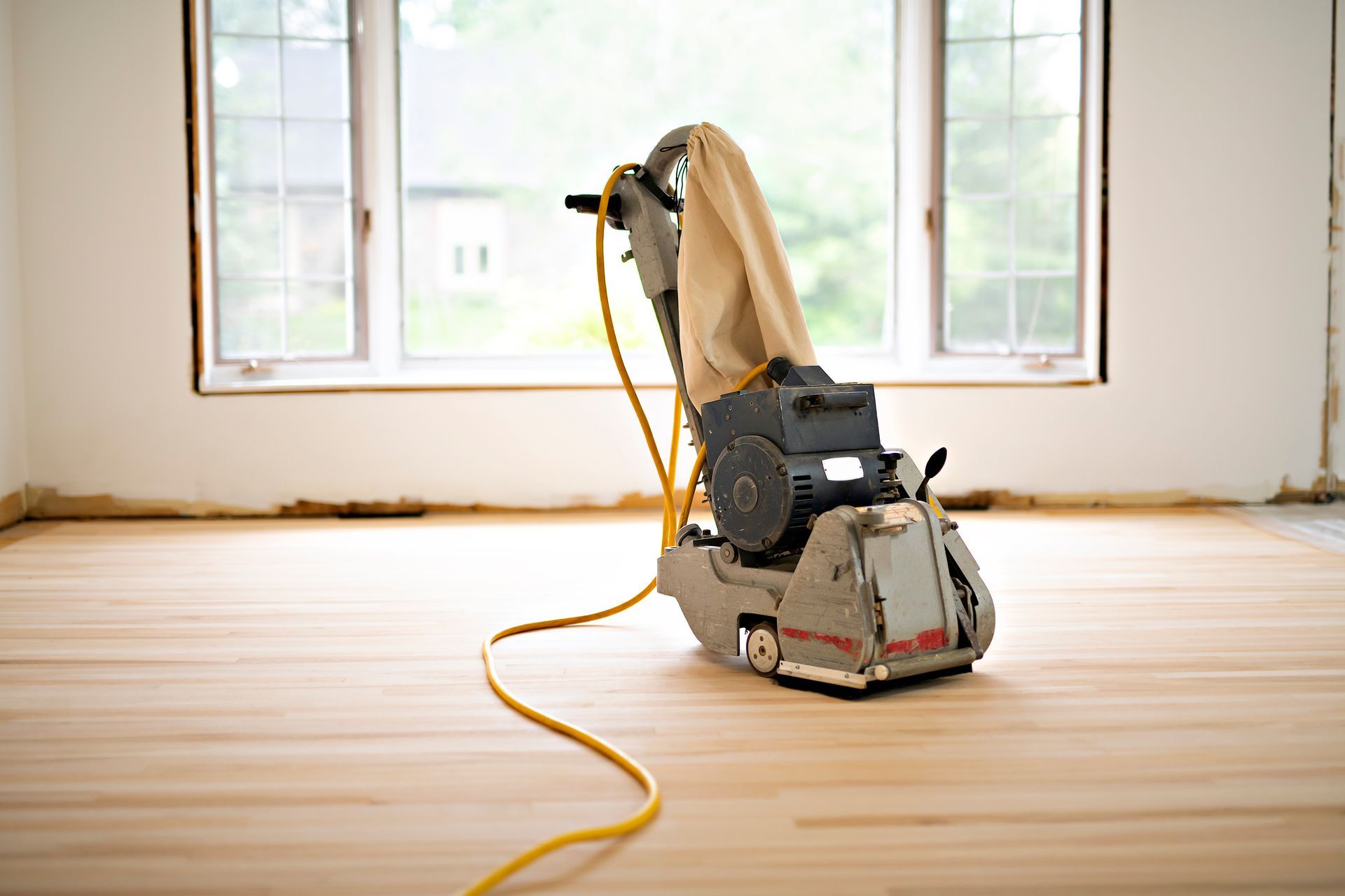 A drum floor sander with a dust bag sits in the center of an empty room with light wood floors and a large bay window.