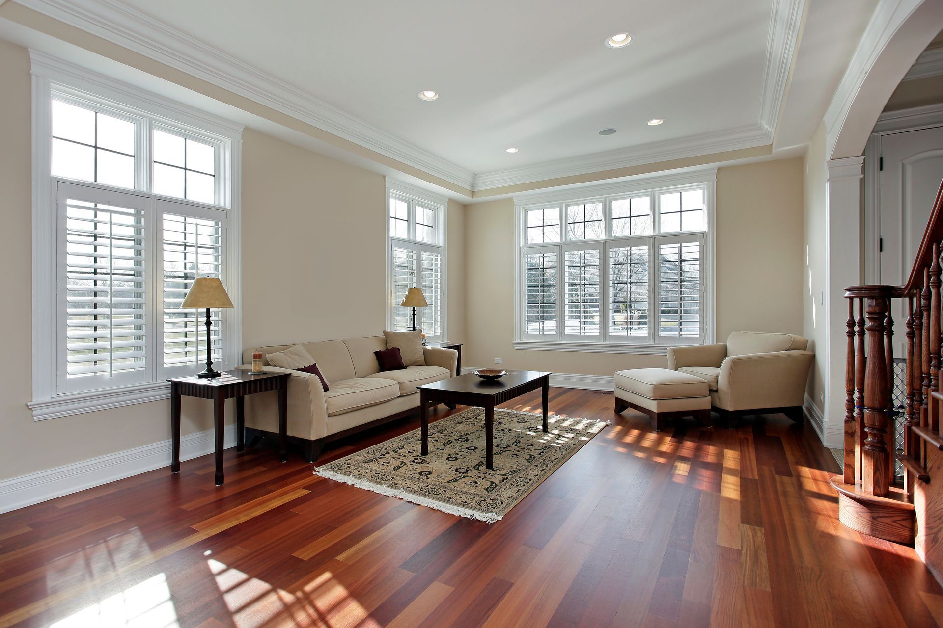 A bright living room with cream walls, hardwood floors, a beige sofa, two armchairs, and windows with white shutters.