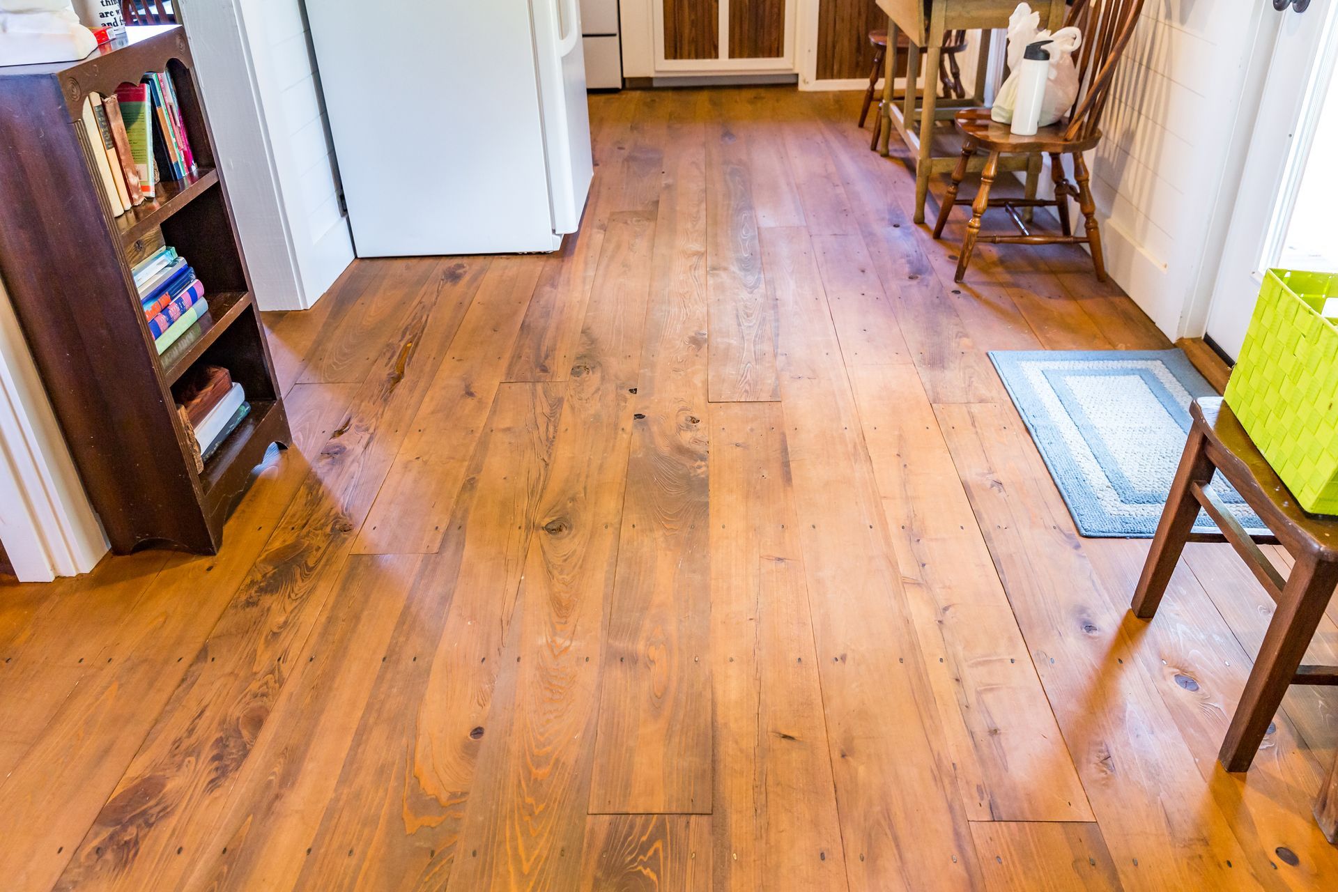 Wide-plank wood floors with a natural finish in a kitchen with a wooden bookshelf and dining chair.