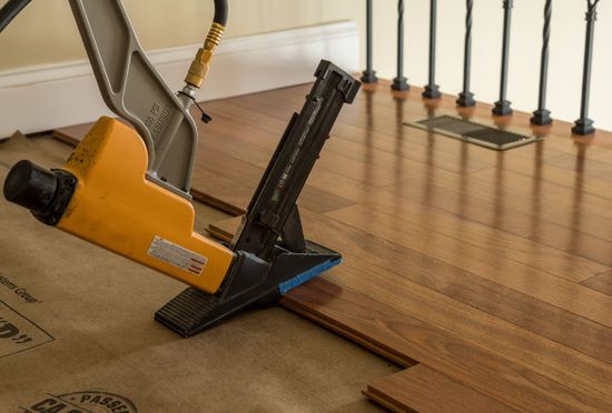 A yellow and black pneumatic flooring nailer being used to install hardwood planks on a floor underlayment.