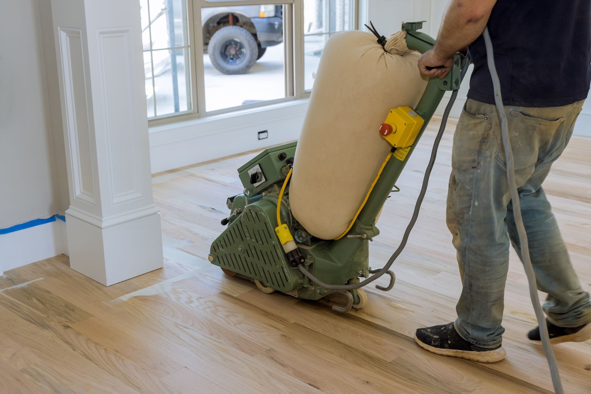 A worker uses a large, green industrial floor sander on hardwood floorboards in a room with white trim.