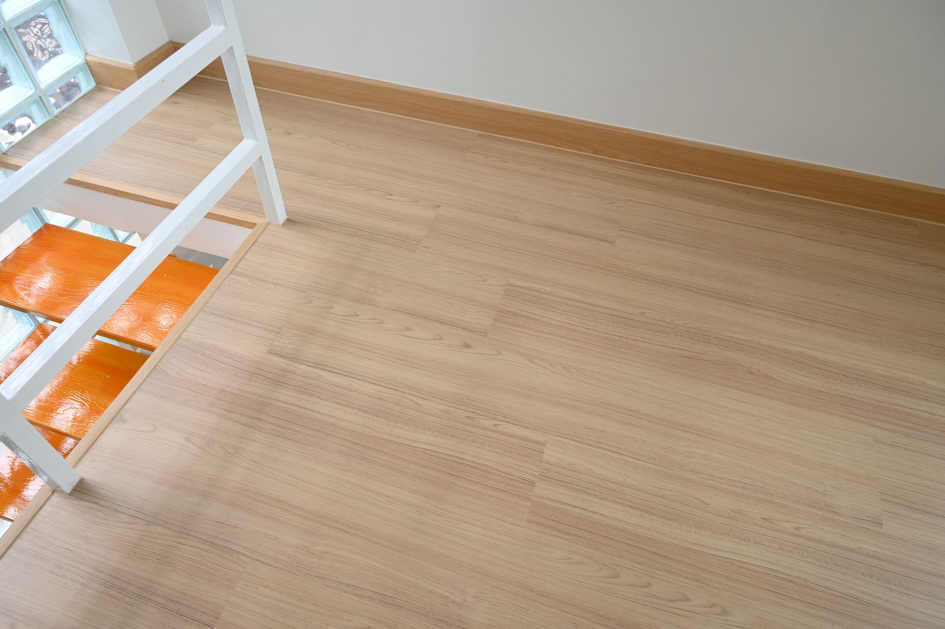 A light wood-grain floor in a room with white railings, a tan baseboard, and a partial view of orange steps.