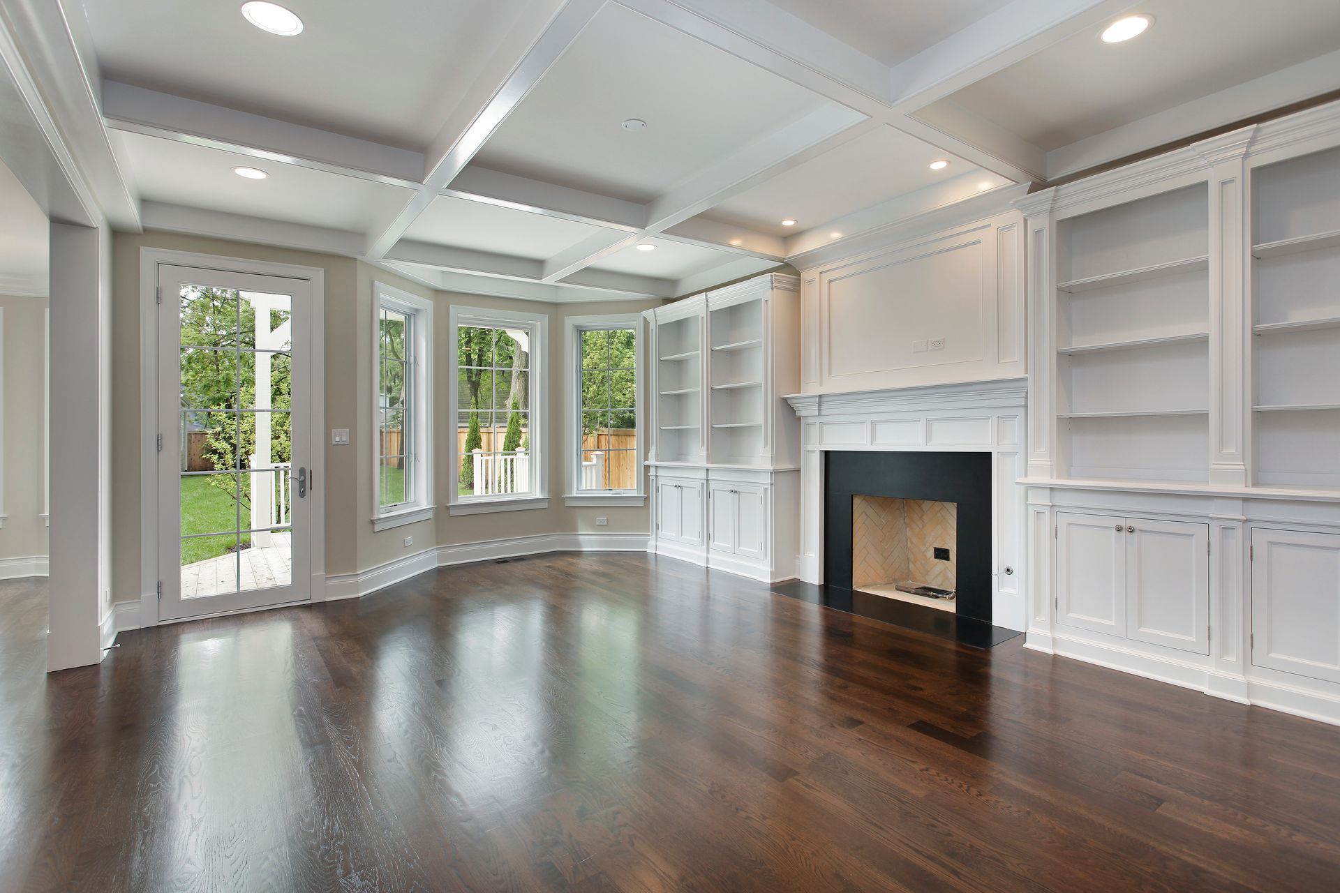 A bright, empty living room with dark hardwood floors, white coffered ceilings, built-in shelving, and a black fireplace.