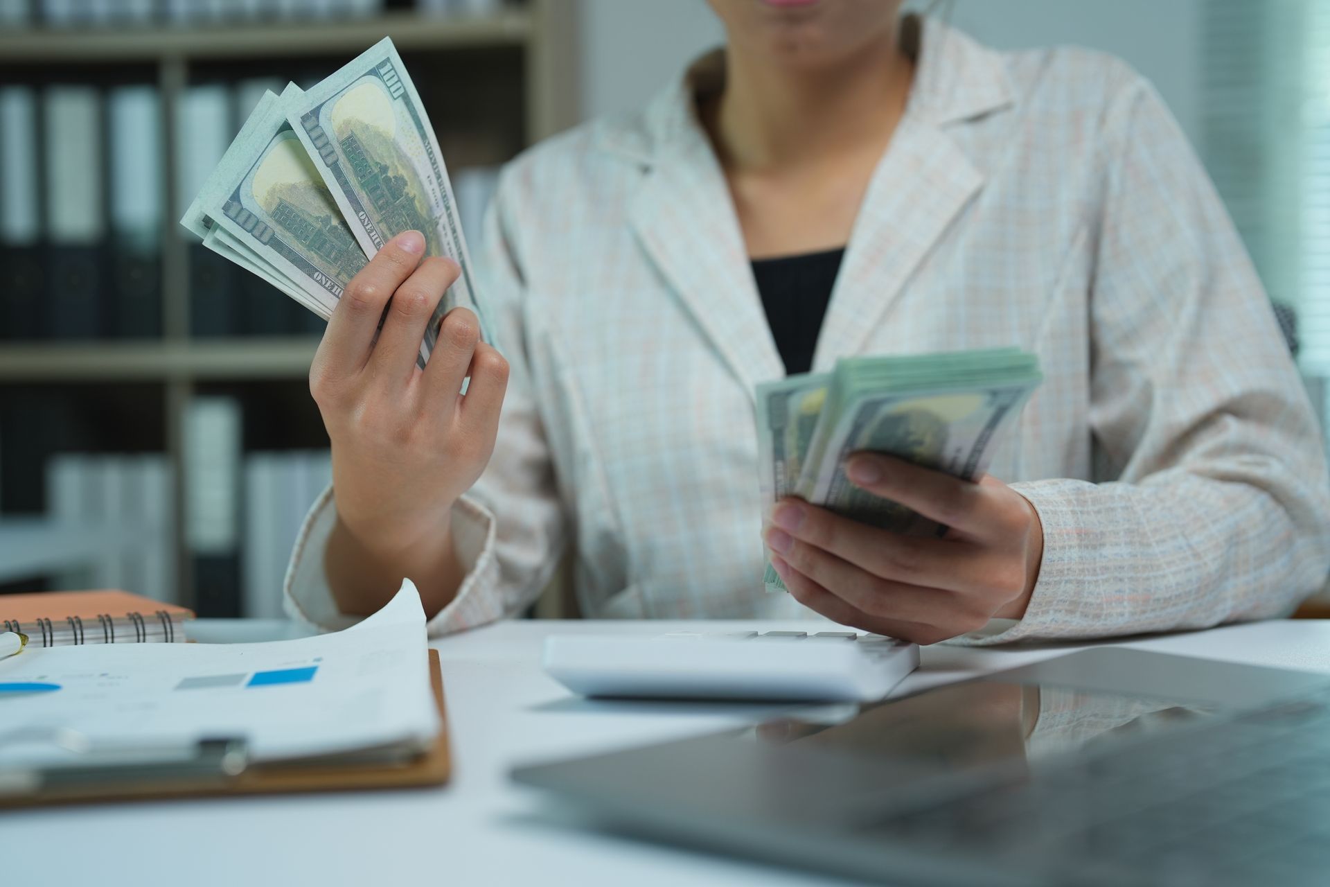 Person counting U.S. dollar bills at a desk with a calculator and financial documents nearby.