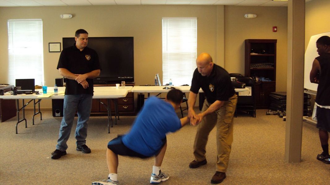 A man in a blue shirt is squatting down while a man in a black shirt stands behind him