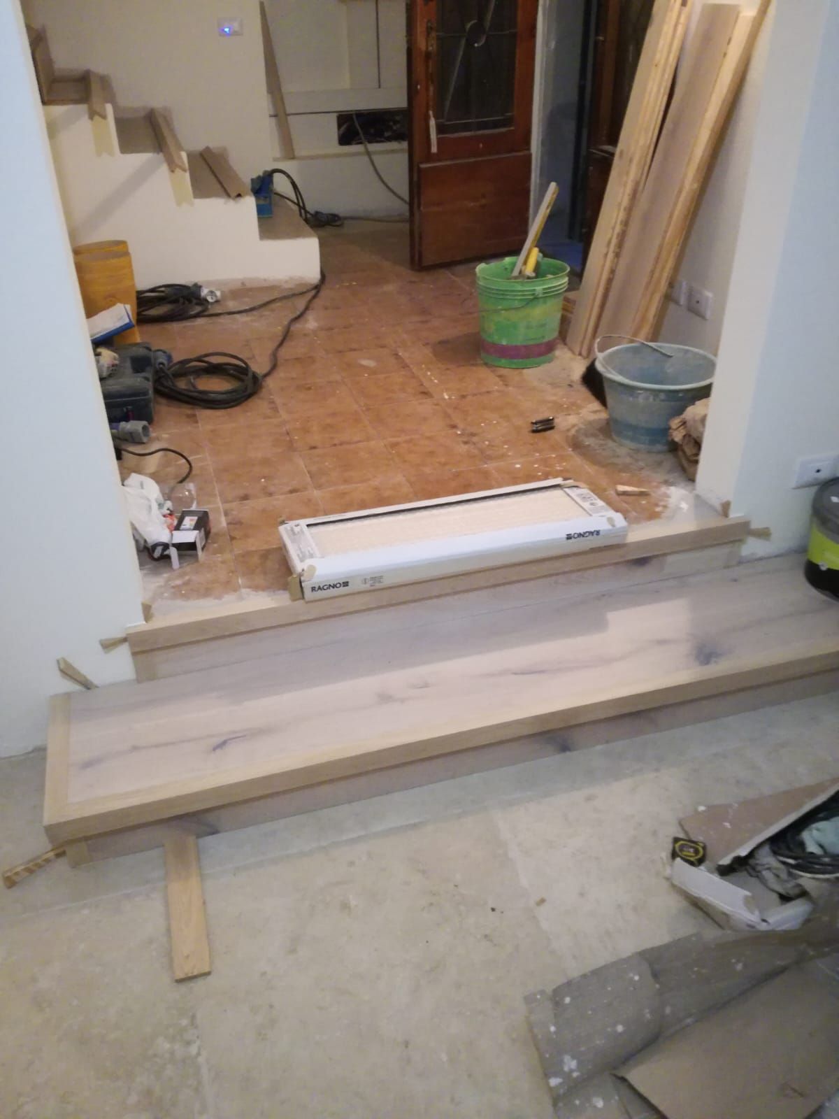 Ingresso interno in ristrutturazione, con assi per la rampa, pavimento piastrellato, attrezzi e un secchio nelle vicinanze