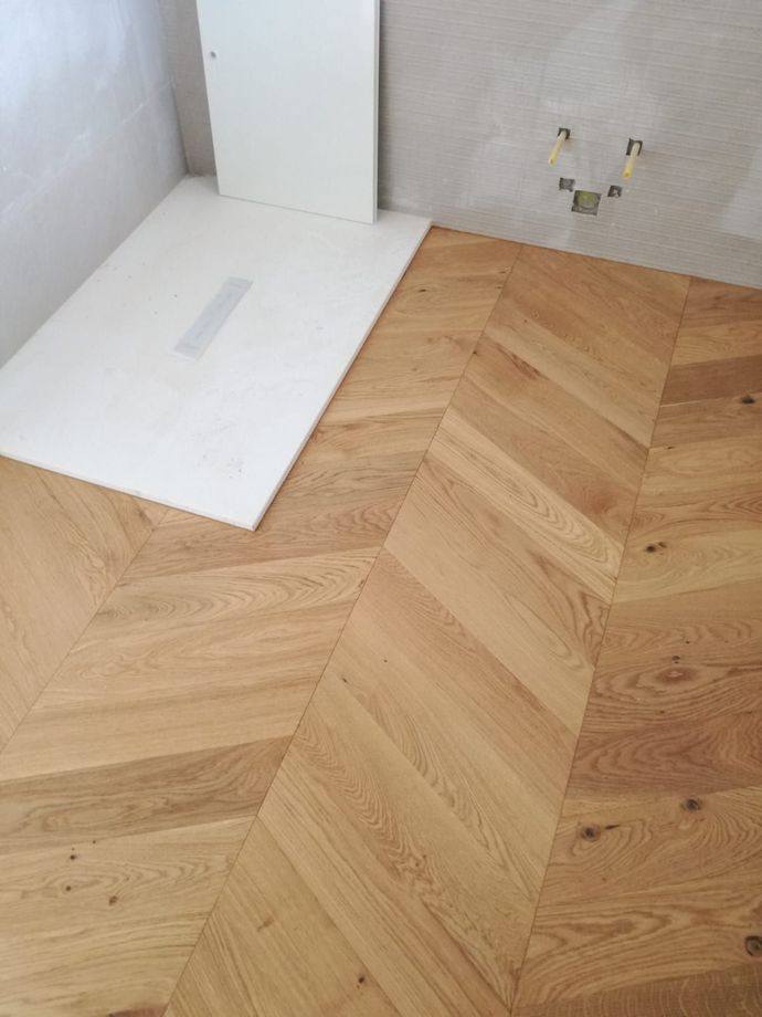 Pavimento in legno a spina di pesce parzialmente posato, accanto a un pannello di sottofondo bianco e a una zona della parete con una presa ancora da rifinire