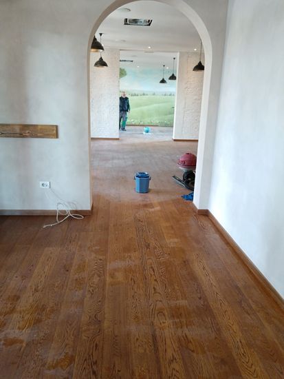 Corridoio con pavimento in legno, pareti bianche, porta ad arco e vista su una stanza con lampade a sospensione e affaccio sull’esterno
