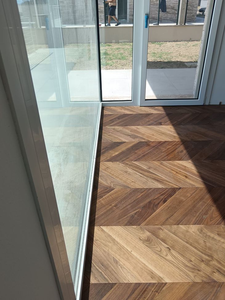 Ingresso luminoso con pavimento in legno a spina di pesce, parete in vetro e porta aperta su un patio esterno.
