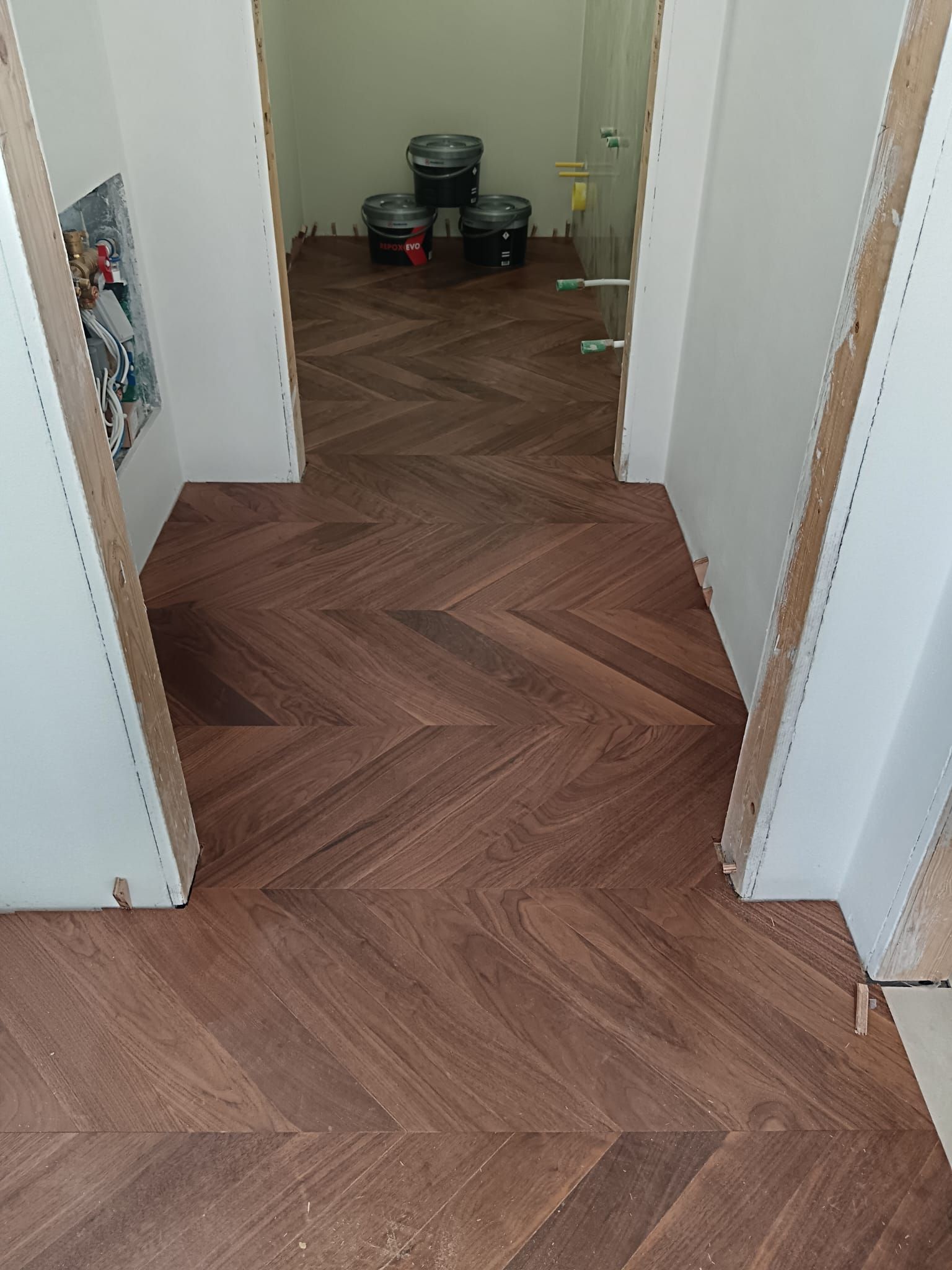 Corridoio con pavimento in legno a spina di pesce che conduce a una stanza con sedie impilate e una pianta in fondo