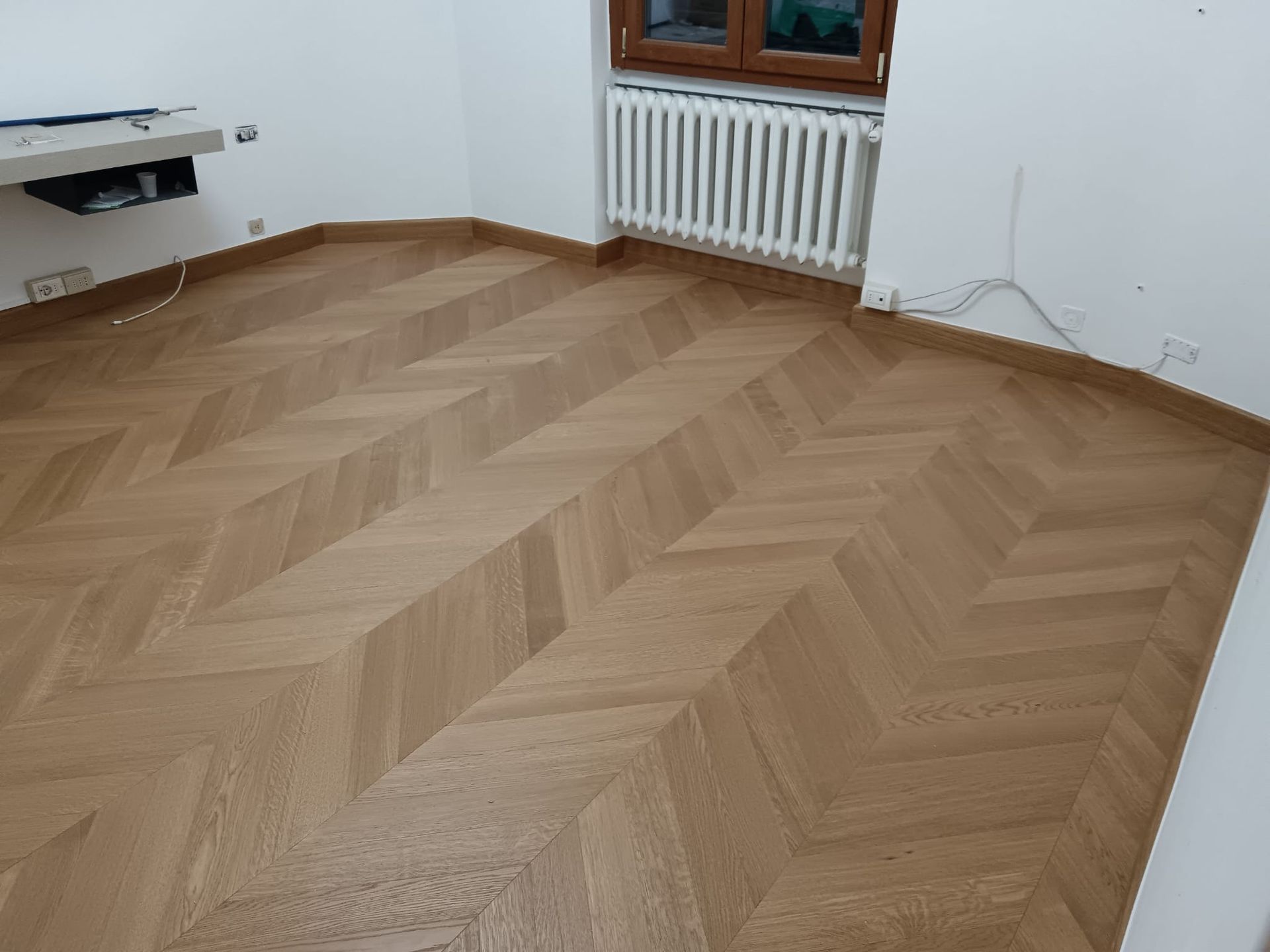 Stanza vuota con pavimento in legno a spina di pesce, pareti bianche e una finestra sopra un termosifone.