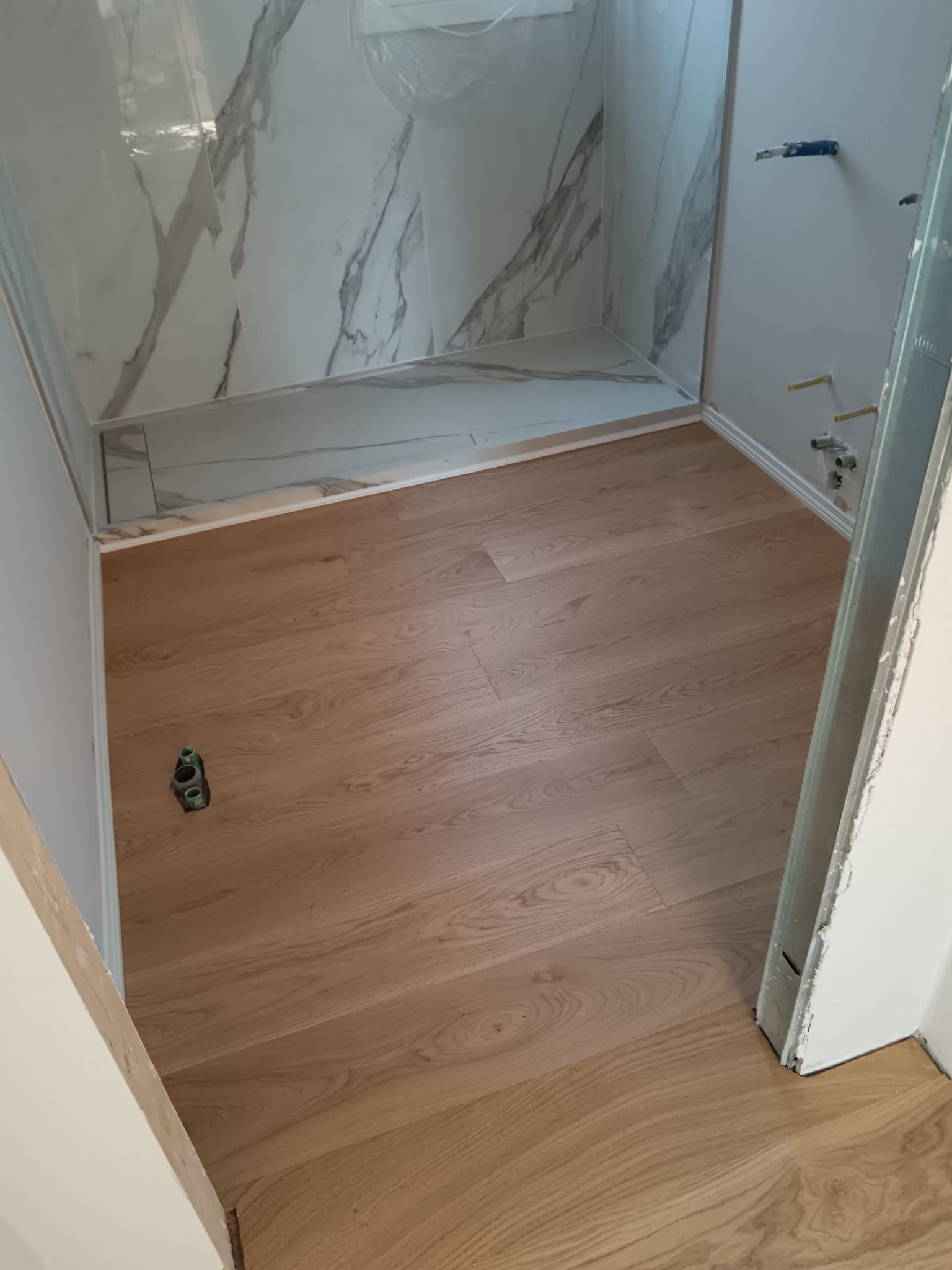 Box doccia vuota con pareti rivestite in piastrelle effetto marmo e pavimento effetto legno, all’interno di un bagno.