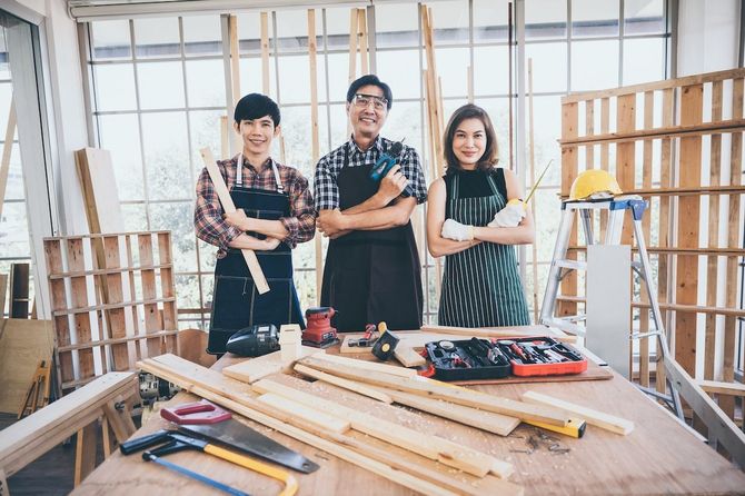 Three people standing in a woodworking workshop behind a table with lumber and tools