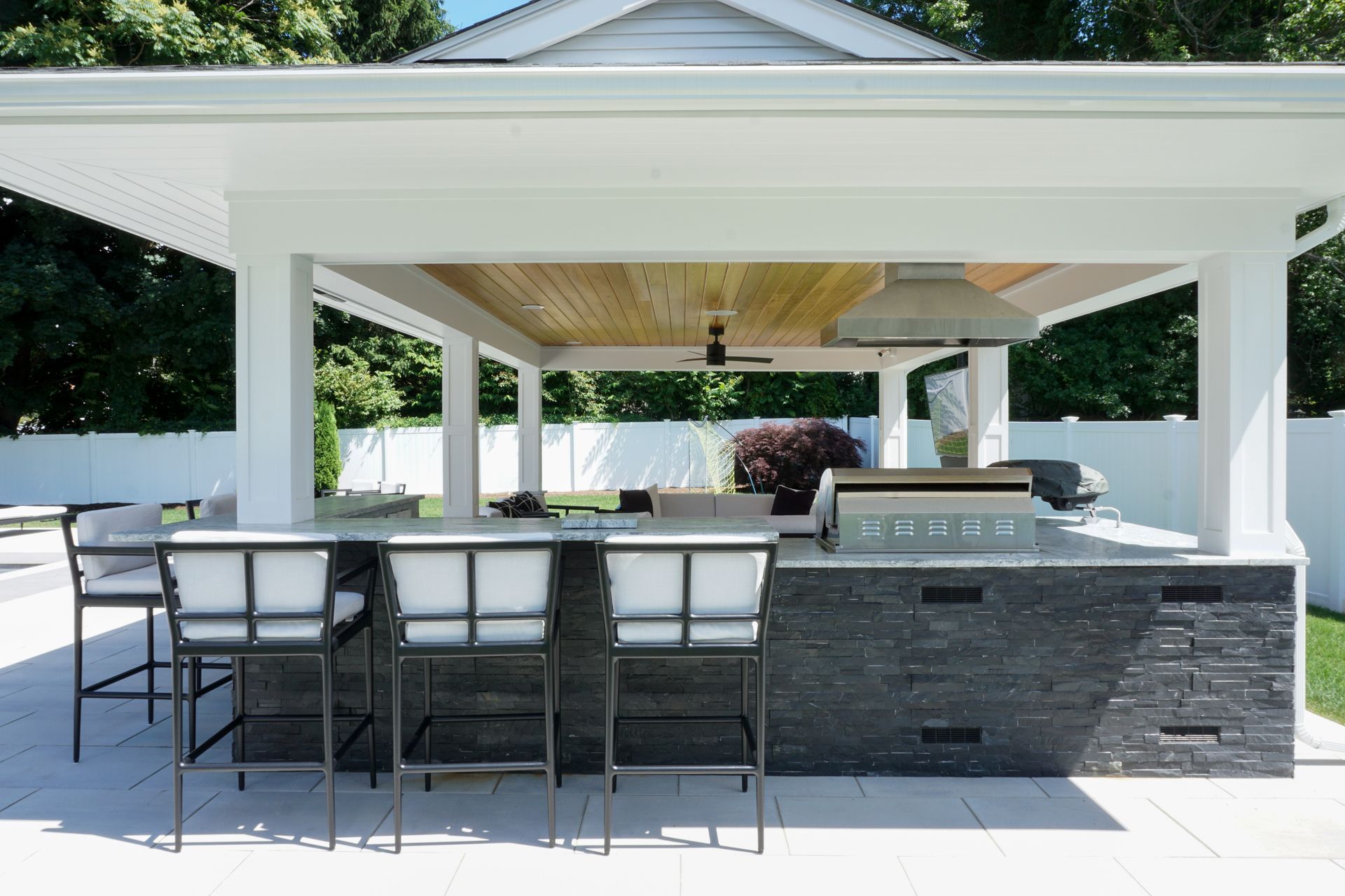 Outdoor patio with bar seating and a stone grill/kitchen under a white pergola, overlooking a pool