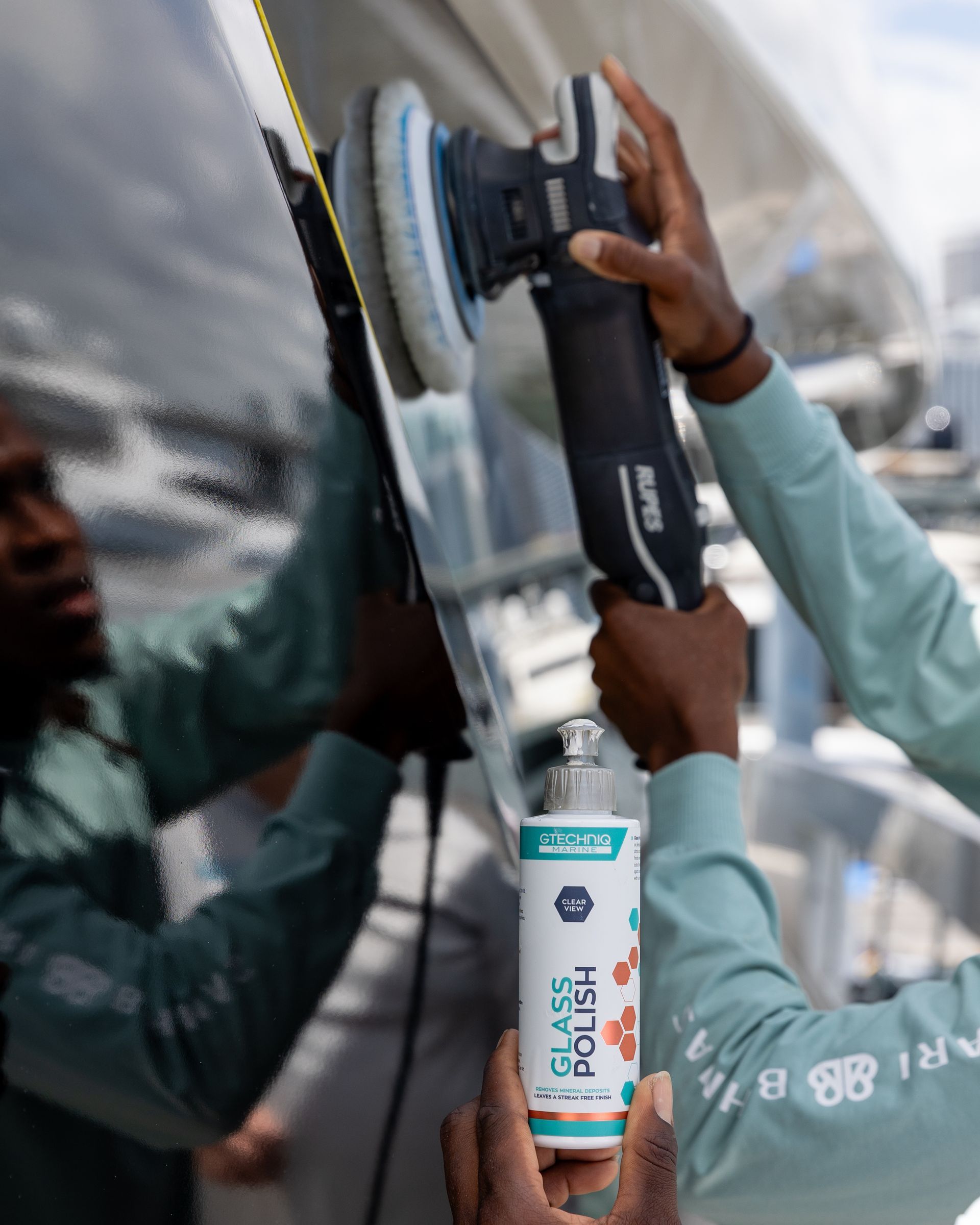 Two workers polishing a vehicle with a handheld buffer near a reflective car surface.