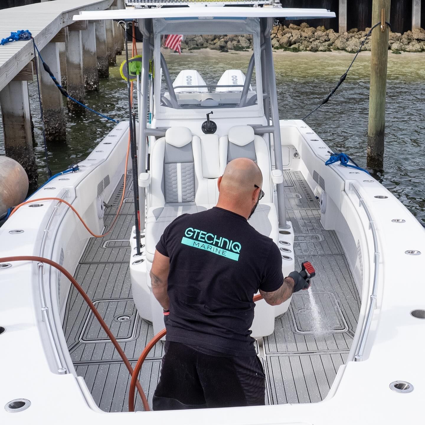 Man washing a white boat deck at a dock, with water and pilings in the background.