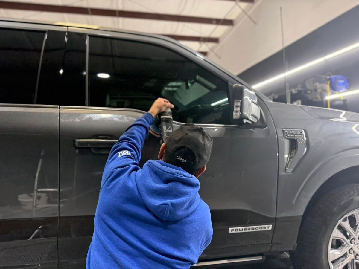 A person in a blue hoodie sprays a car window on a gray pickup truck. The car is in a garage with bright lights.