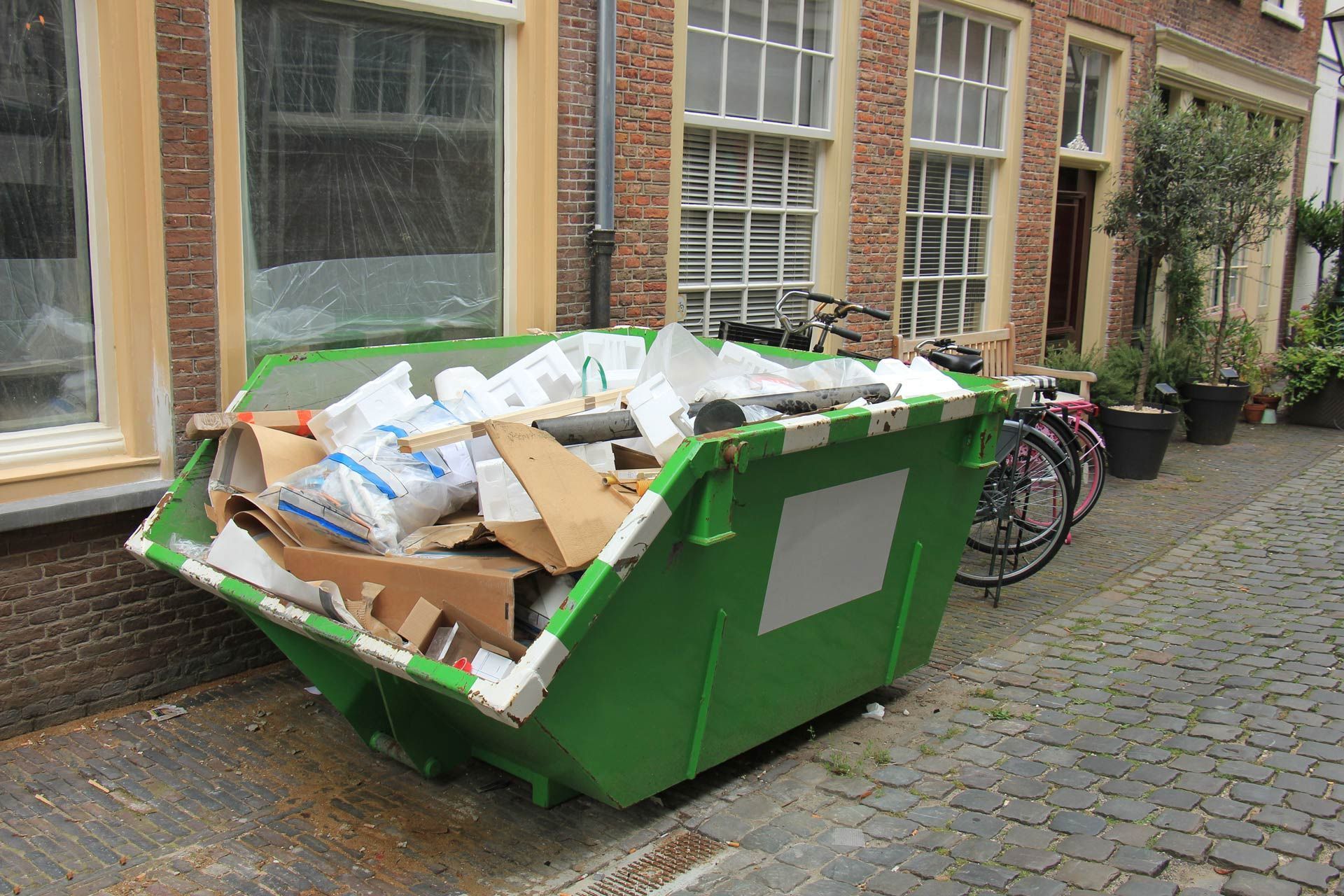 Green dumpster overflowing with trash on a brick-lined street, near a building with windows.