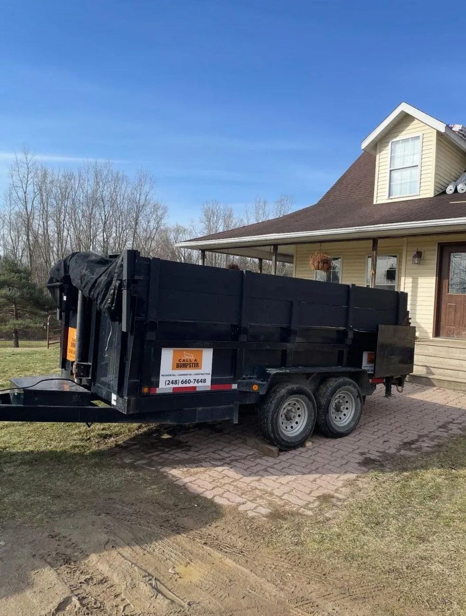 Black dump trailer parked on gravel near a house, blue sky above.