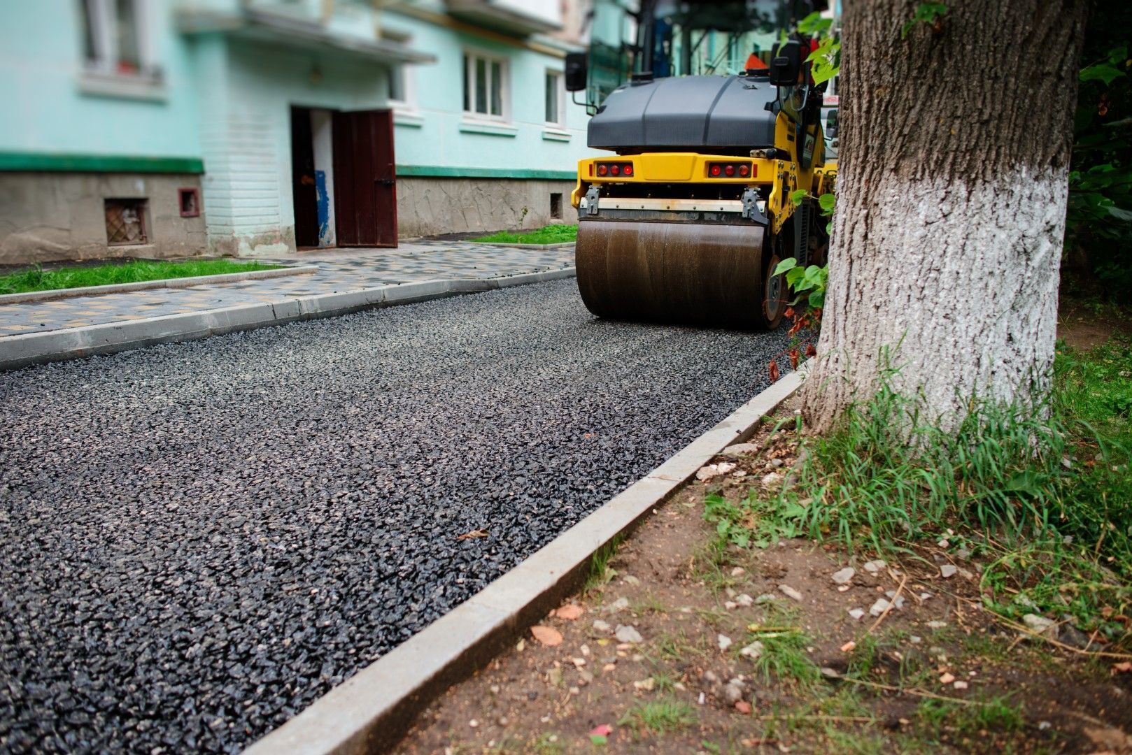 A yellow roller is rolling gravel on the side of a road.