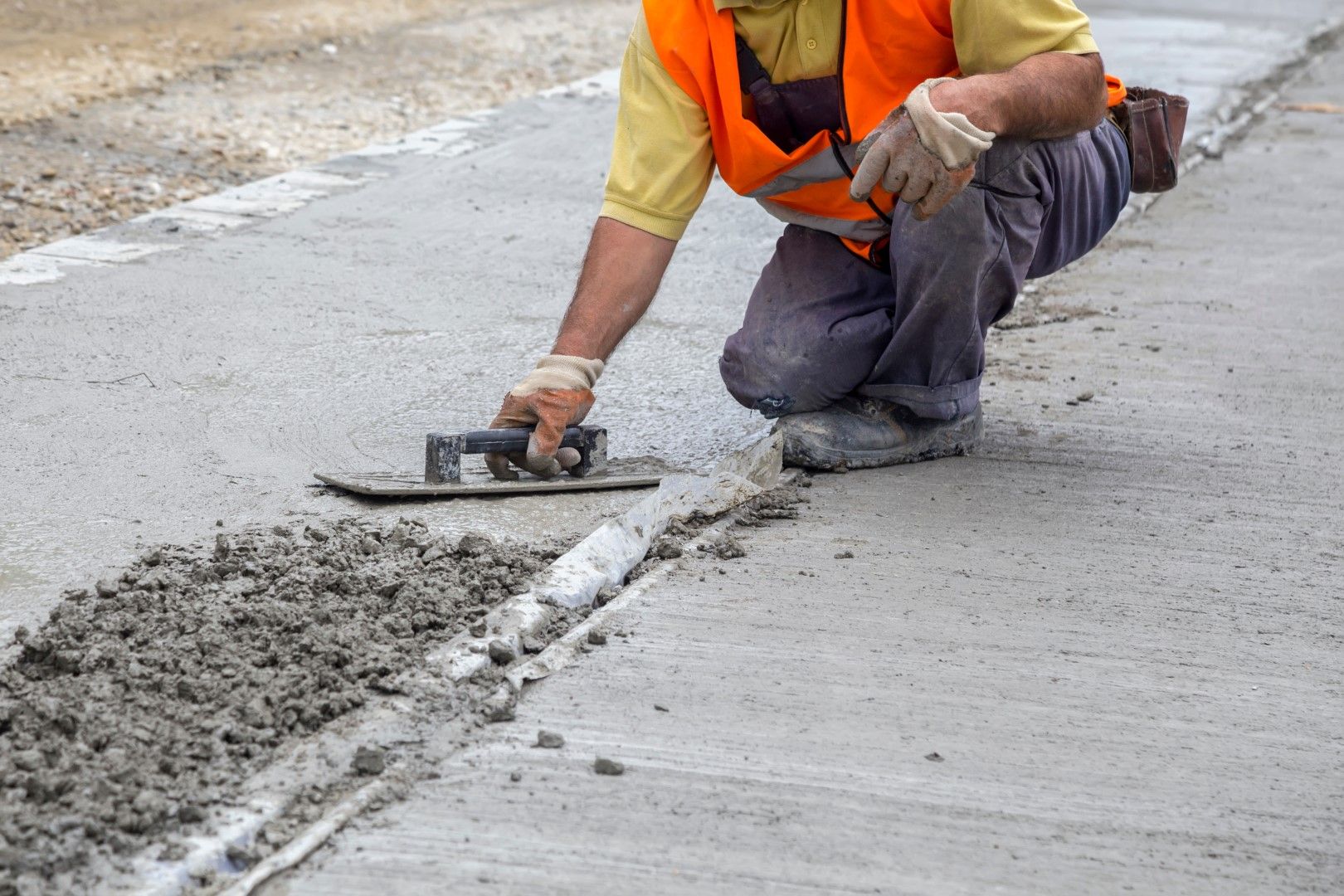A construction worker is kneeling down and spreading concrete on a sidewalk.
