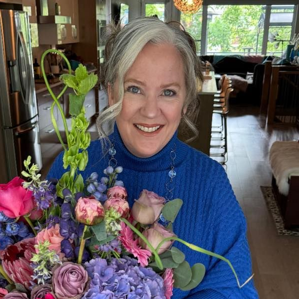 Woman smiling indoors, holding a bouquet of pink and purple flowers in a cozy home setting