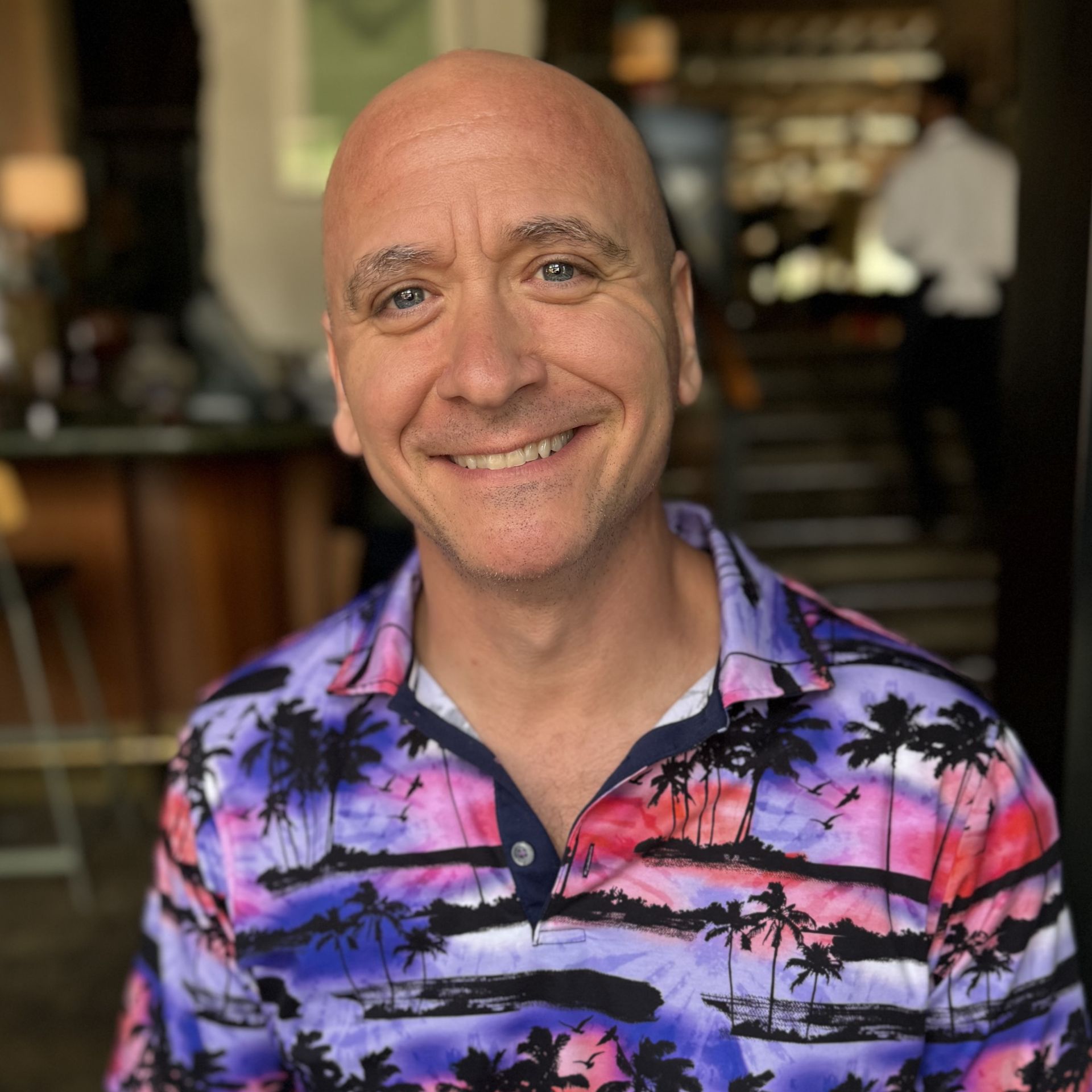 A bald man wearing a colorful shirt with palm trees on it is smiling for the camera.
