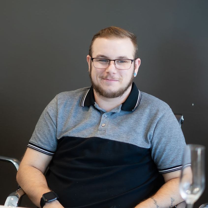 Man with glasses and a gray and black polo shirt smiles, seated near a table.