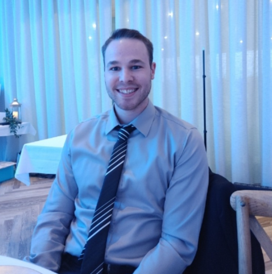 Man with light complexion smiling, wearing grey shirt and striped tie, seated at a table.