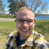 Smiling person in glasses and yellow plaid shirt outdoors by a lake on a sunny day