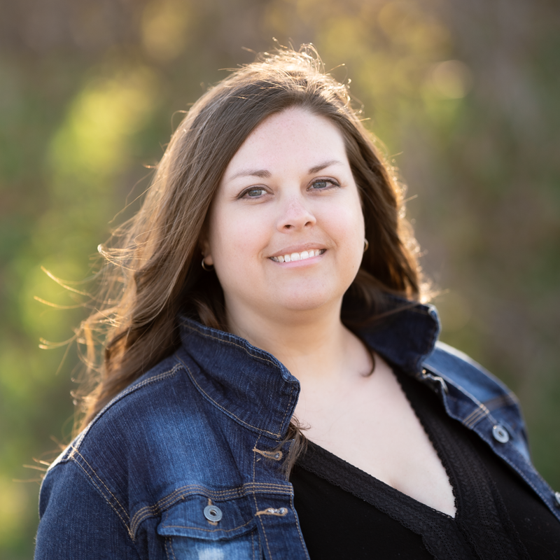 Woman smiling outdoors in a denim jacket against a blurred green background