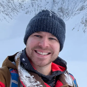 Smiling person in a dark knit beanie and snowy jacket against a snowy mountain backdrop