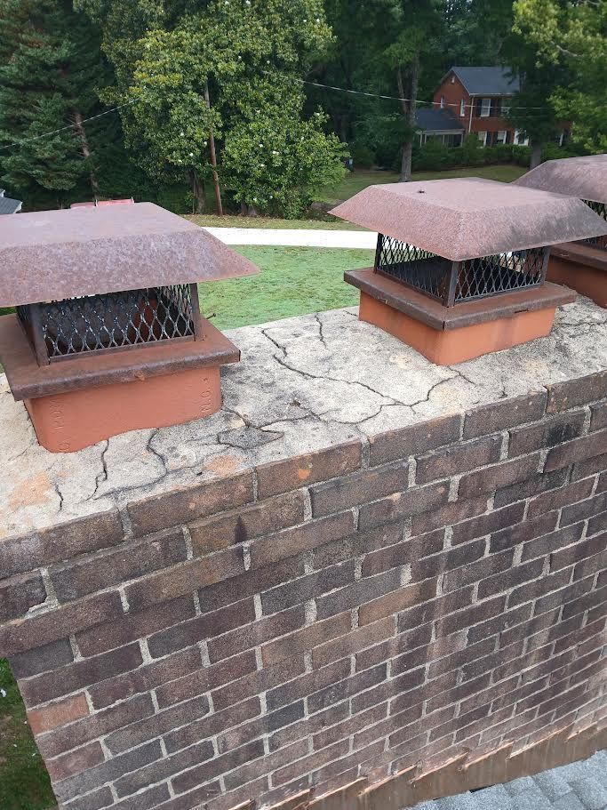 Two brick chimney pots on a rooftop, with trees and a house in the background.