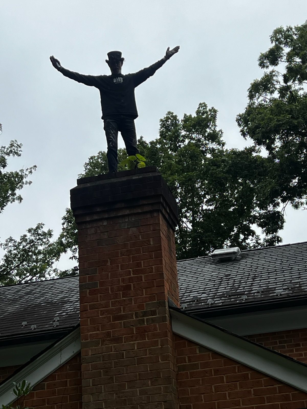 Statue with arms raised atop a brick chimney above a house roof, framed by trees and cloudy sky