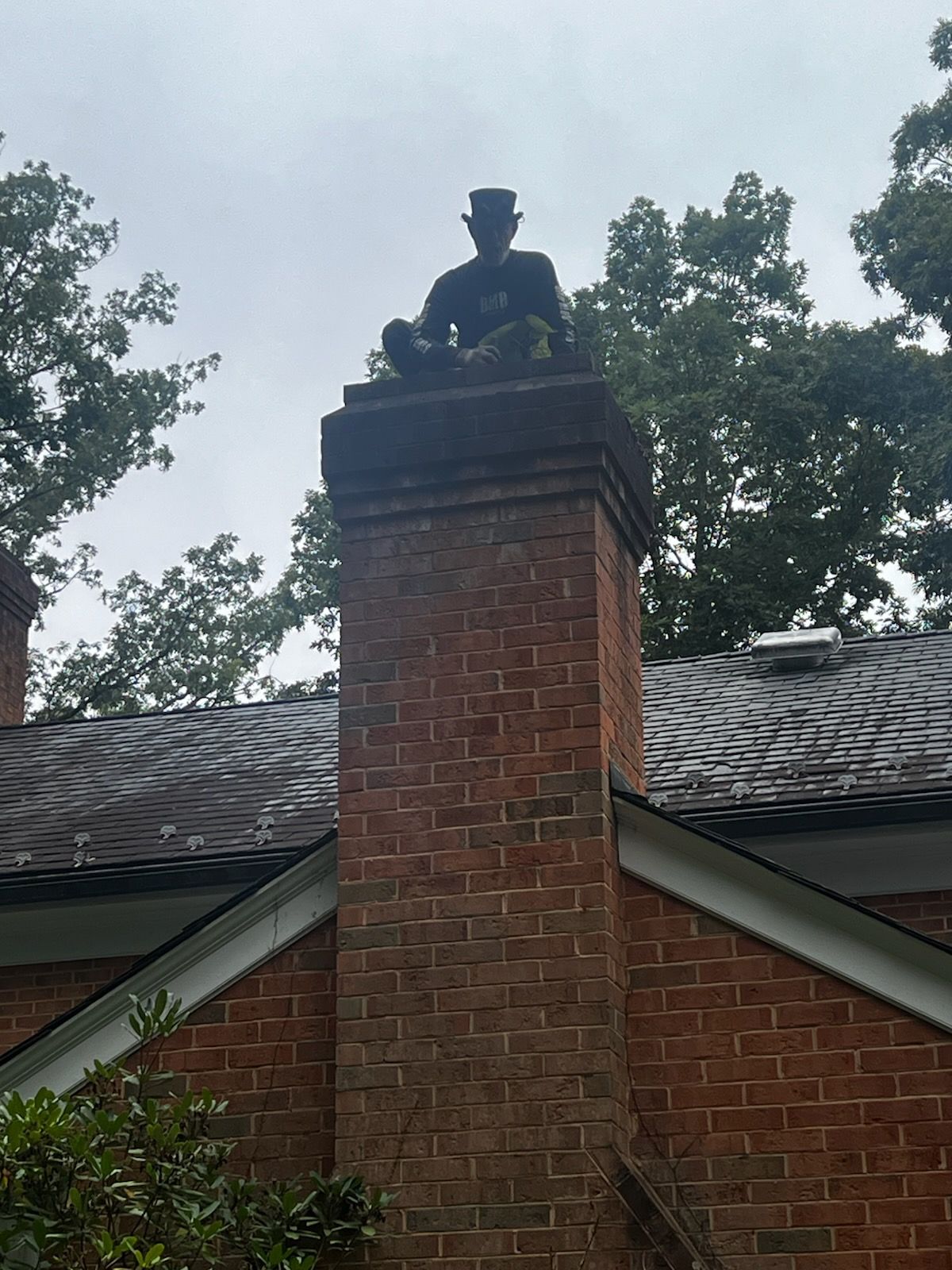 Statue of a seated man atop a brick chimney on a house, framed by trees and rooftop shingles