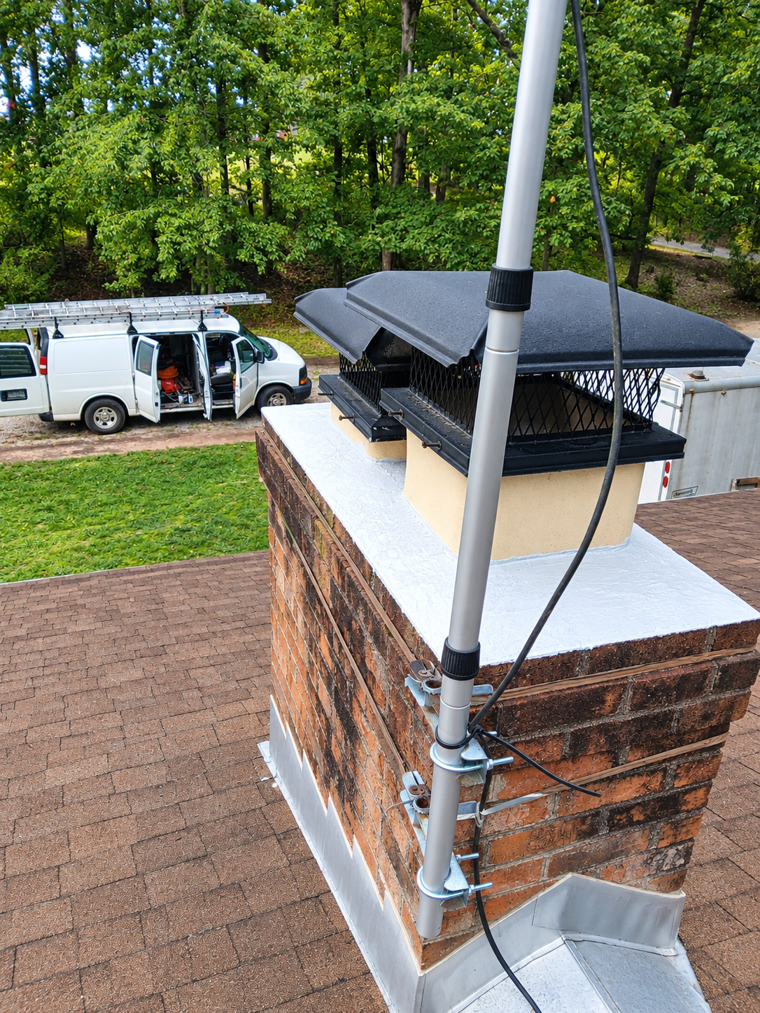Roof edge with white van parked below, seen from a brick chimney under green trees.
