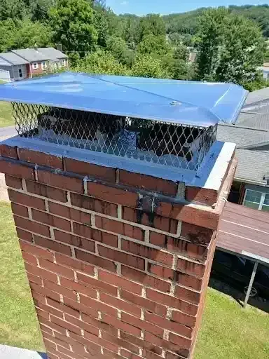 Blue chimney cap on a brick chimney with metal mesh, viewed outdoors above a grassy yard.
