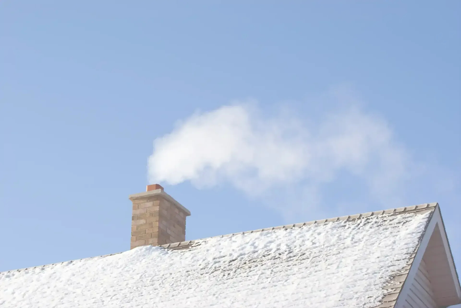Snow-covered roof with a stone chimney releasing smoke against a blue sky