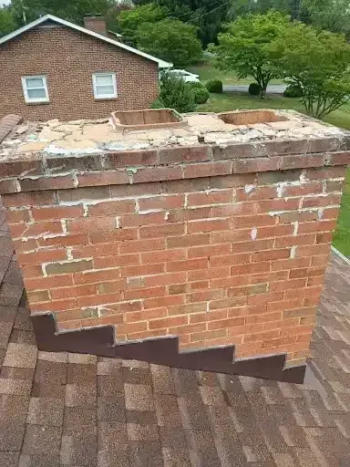 Damaged brick chimney with missing top bricks on a shingled roof, with a house and trees in the background