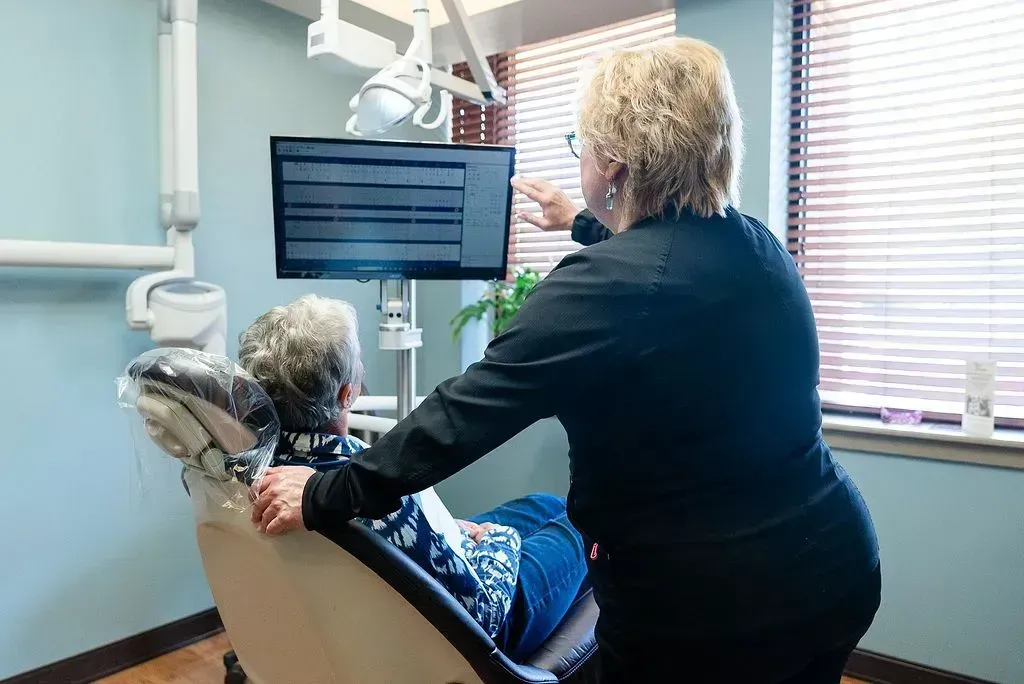 Dental hygienist assisting patient in a dental chair beside a monitor and window blinds