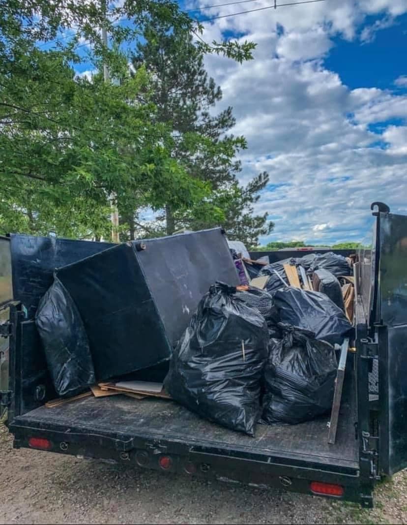 Truck bed filled with black trash bags and furniture beside trees under a cloudy sky