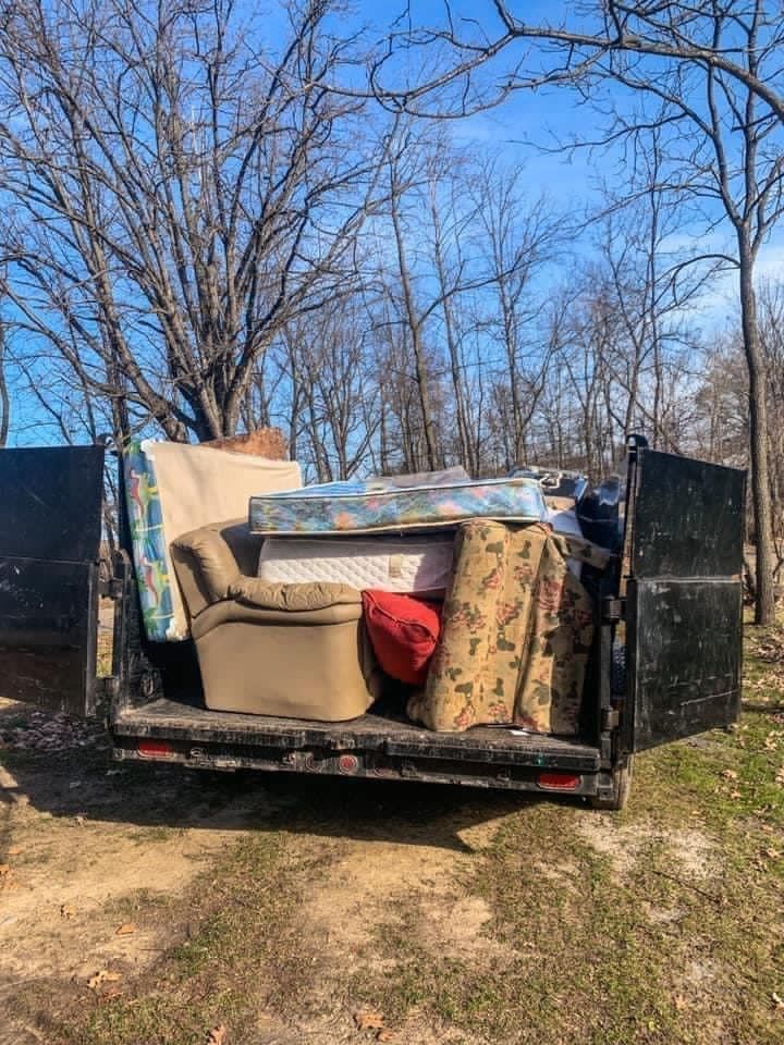 Dump trailer loaded with furniture and a rug in a wooded yard on a sunny day