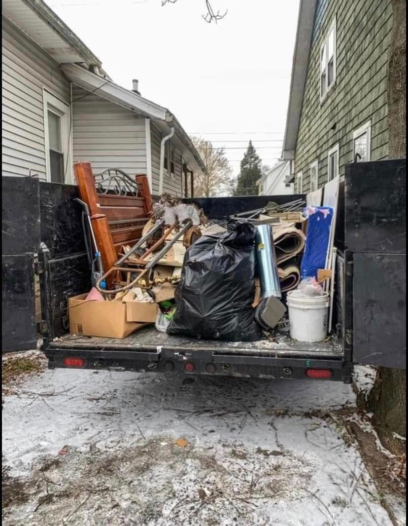 Loaded trailer with mixed junk between houses on a snowy driveway.