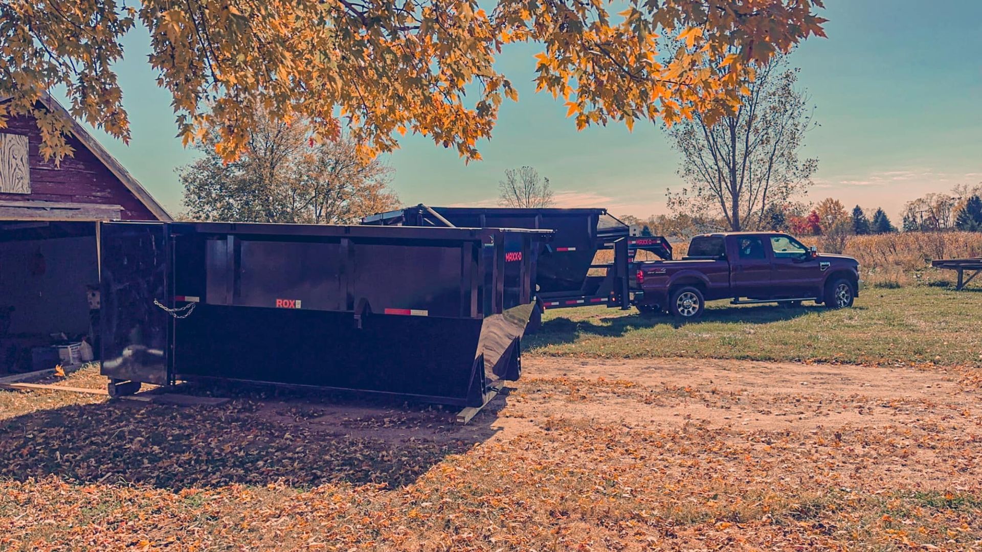 Yard with a black dumpster and pickup trucks under a tree beside a house on a sunny day